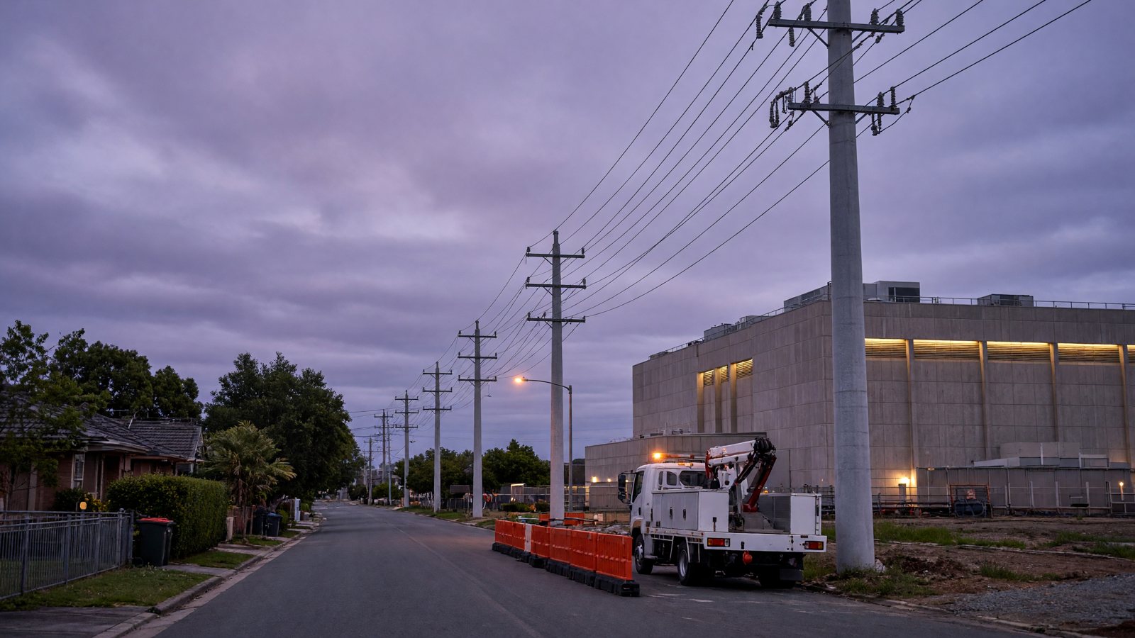 Illustration: tall concrete power poles and construction barriers on a suburban street at dusk, with a data centre in the background.
